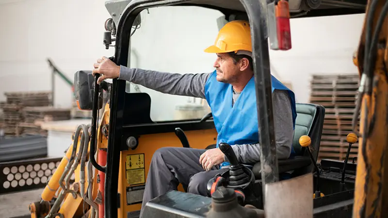 A person wearing a yellow hard hat and a blue safety vest is operating a piece of heavy machinery