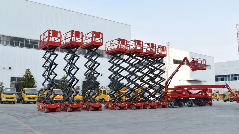 Industrial yard displaying various red scissor lifts, illustrating types of scissor lifts, with yellow trucks and factory structures in the background, along with a red articulating boom lift on the right.